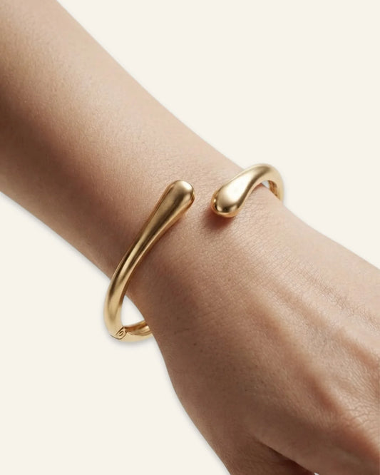 Duo Curve Bracelet, a gold-tone open cuff bracelet with smooth, teardrop-shaped terminals and a hinged closure, worn on a wrist against a neutral background.