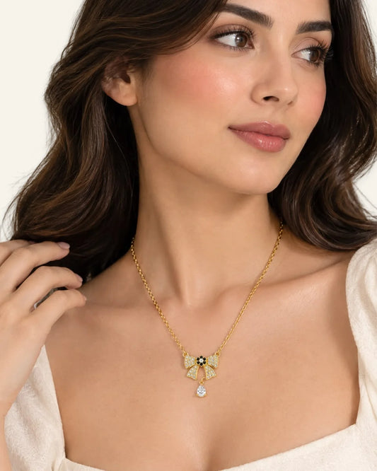 Close-up of a brunette model wearing a yellow gold ribbon bow pendant necklace with pavé-set crystals and a dangling pear-shaped stone, styled with an off-white top.