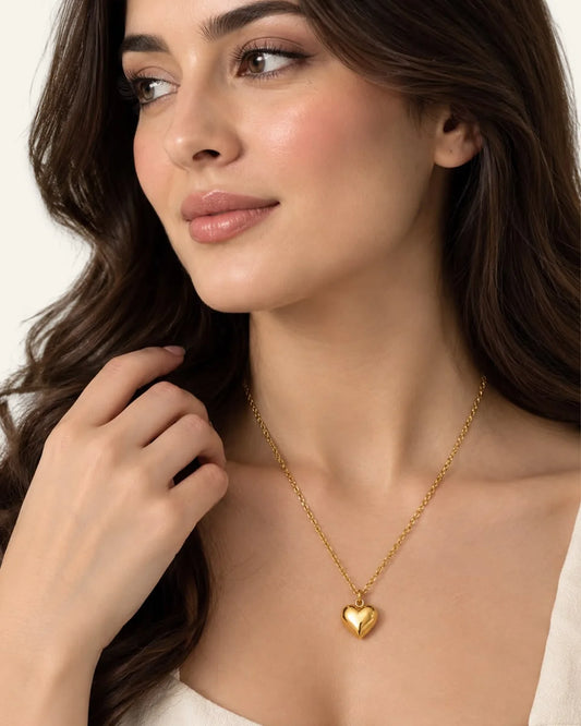 Close-up of a model with dark wavy hair wearing a delicate gold chain necklace with a shiny puffed heart pendant.