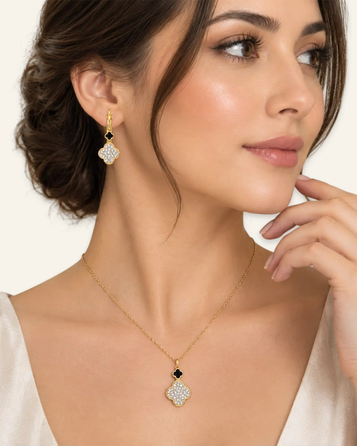 A close-up of a woman wearing a gold necklace and matching drop earring. The jewelry features a gold four-petal pendant and earring set with small black stones and clear pavé-style stones, shown against a light cream background.