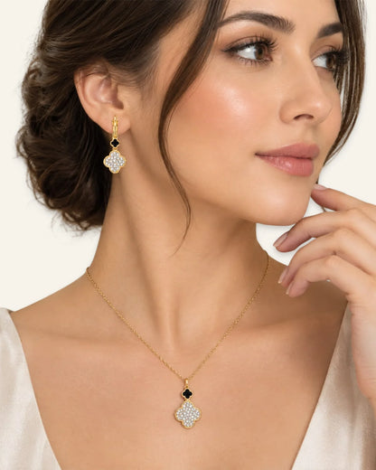 A close-up of a woman wearing a gold necklace and matching drop earring. The jewelry features a gold four-petal pendant and earring set with small black stones and clear pavé-style stones, shown against a light cream background.