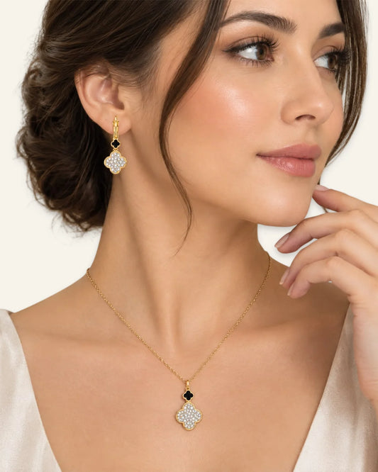 A close-up of a woman wearing a gold necklace and matching drop earring. The jewelry features a gold four-petal pendant and earring set with small black stones and clear pavé-style stones, shown against a light cream background.