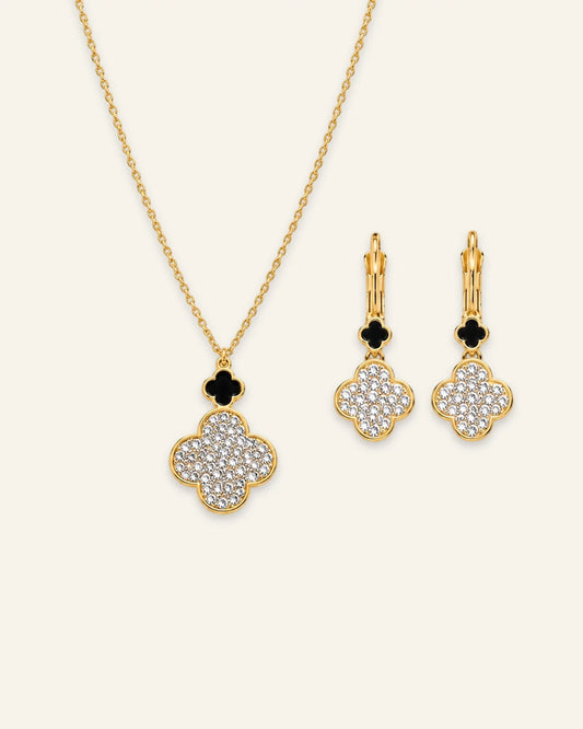 Gold jewelry set featuring a necklace and matching earrings with clover-shaped pendants and floral drop charms, set against a light cream background.