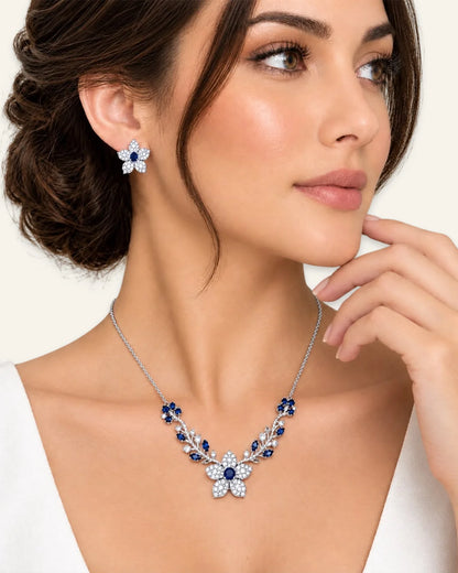 Model wearing a silver floral necklace with sapphire blue center stones and leaf vine accents, paired with matching flower stud earrings.