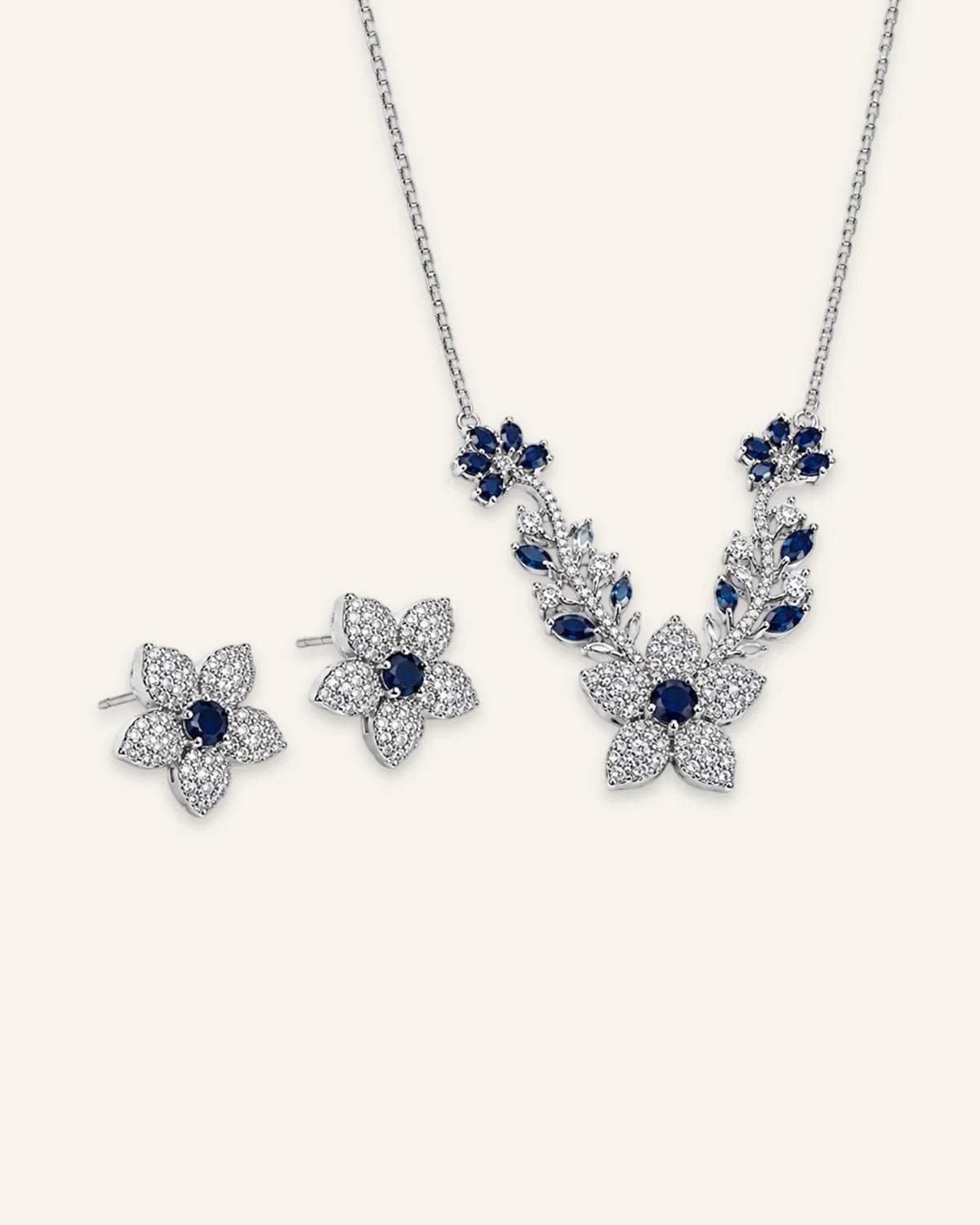 Silver floral necklace and matching stud earrings set featuring sapphire blue center stones, pave-set clear crystal petals, and blue leaf vine accents.