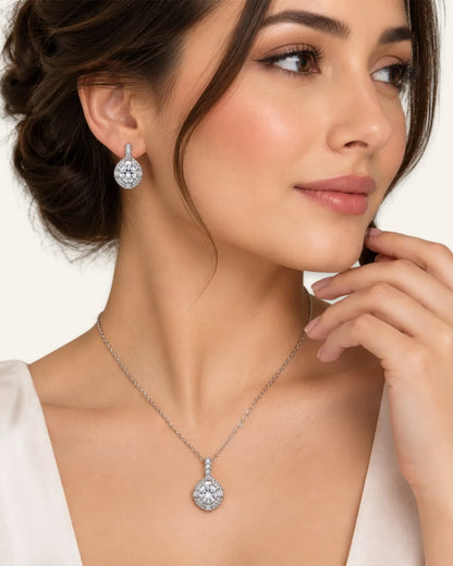 Model wearing a silver halo pendant necklace with a round brilliant clear gemstone and matching silver halo drop earrings.