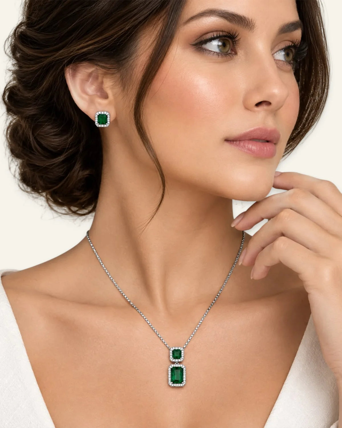A woman wearing a silver necklace with a rectangular green emerald pendant and matching green emerald stud earrings, shown in a close-up portrait against a light neutral background.