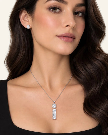 A woman wearing a silver pendant necklace with a vertical baguette-cut clear gemstone and a matching stud earring, shown against a light beige background.