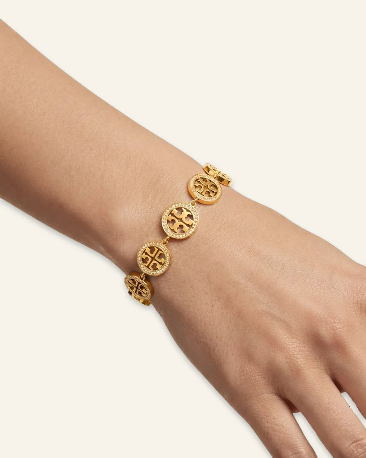 A gold celeste medallion bracelet with interlocking circular logo charms, shown worn on a person's wrist against a warm off-white background.