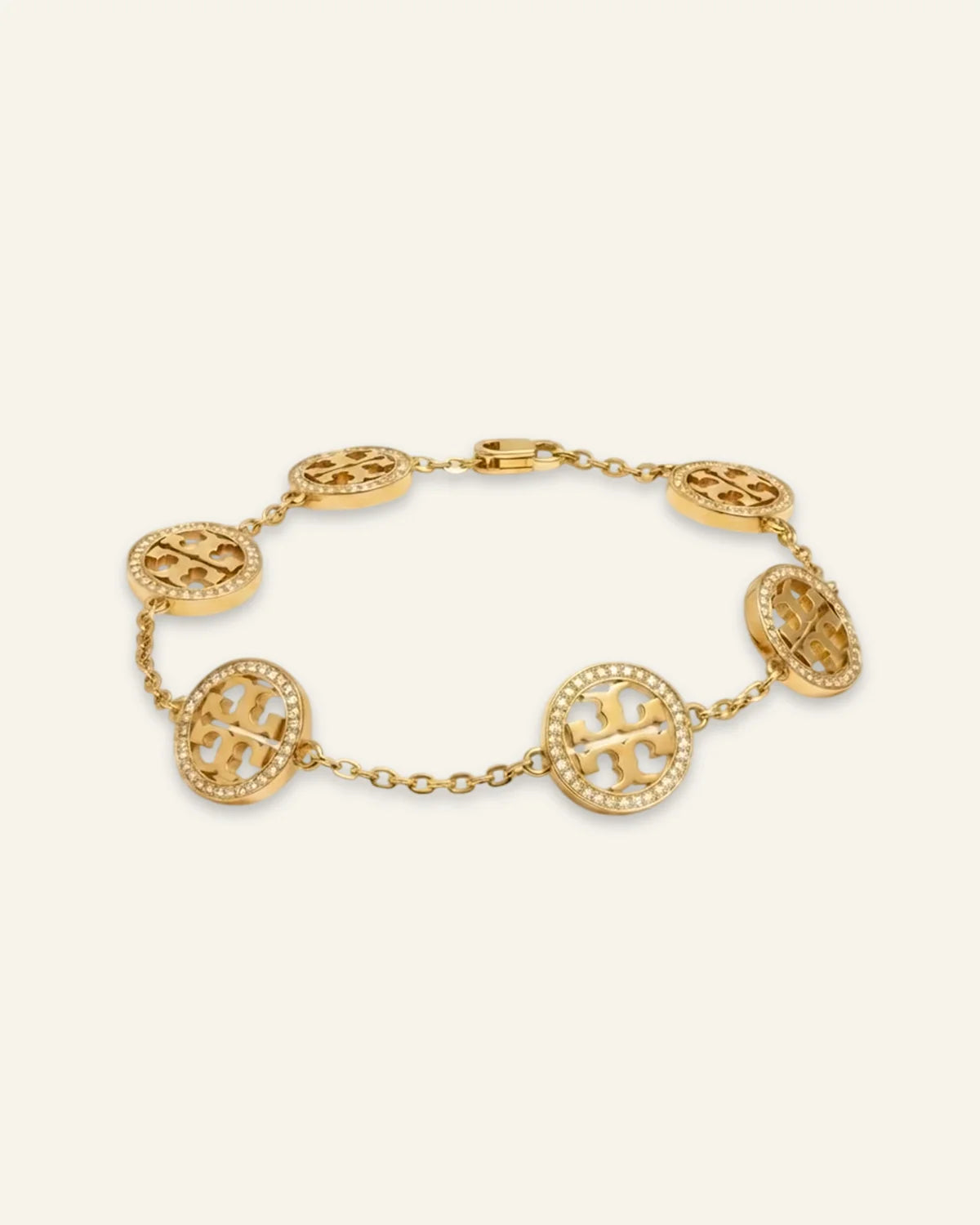 A gold celeste medallion bracelet featuring oval links, each adorned with a raised monogram logo and surrounded by small clear stones, displayed on a light cream background.