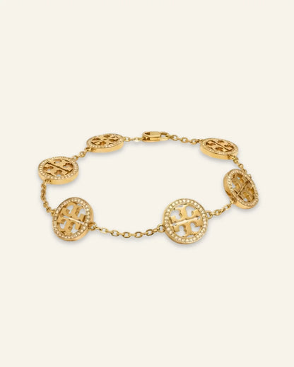 A gold celeste medallion bracelet featuring oval links, each adorned with a raised monogram logo and surrounded by small clear stones, displayed on a light cream background.