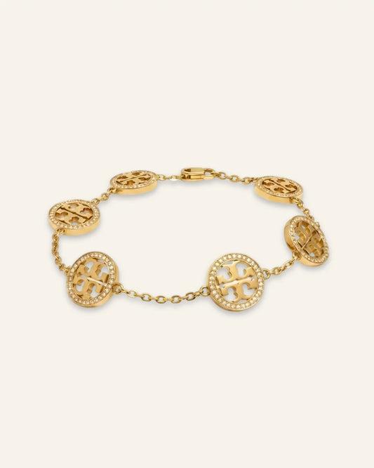 A gold celeste medallion bracelet featuring oval links, each adorned with a raised monogram logo and surrounded by small clear stones, displayed on a light cream background.