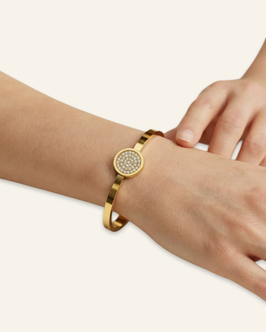 The Celeste Sparkle Bracelet worn on a wrist, featuring a polished gold-tone metal band and a circular centerpiece of shining pave stones against a minimalist neutral background.