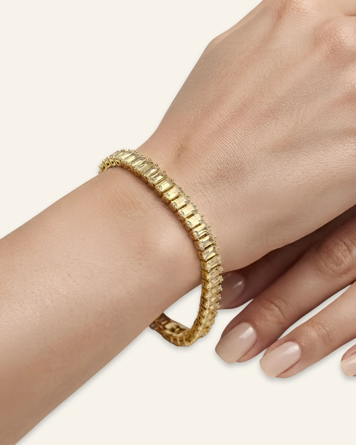 A close-up of a woman's wrist and hand wearing a gold dazzling baguette cut  tennis bracelet with rectangular clear stones, set against a solid light beige background.