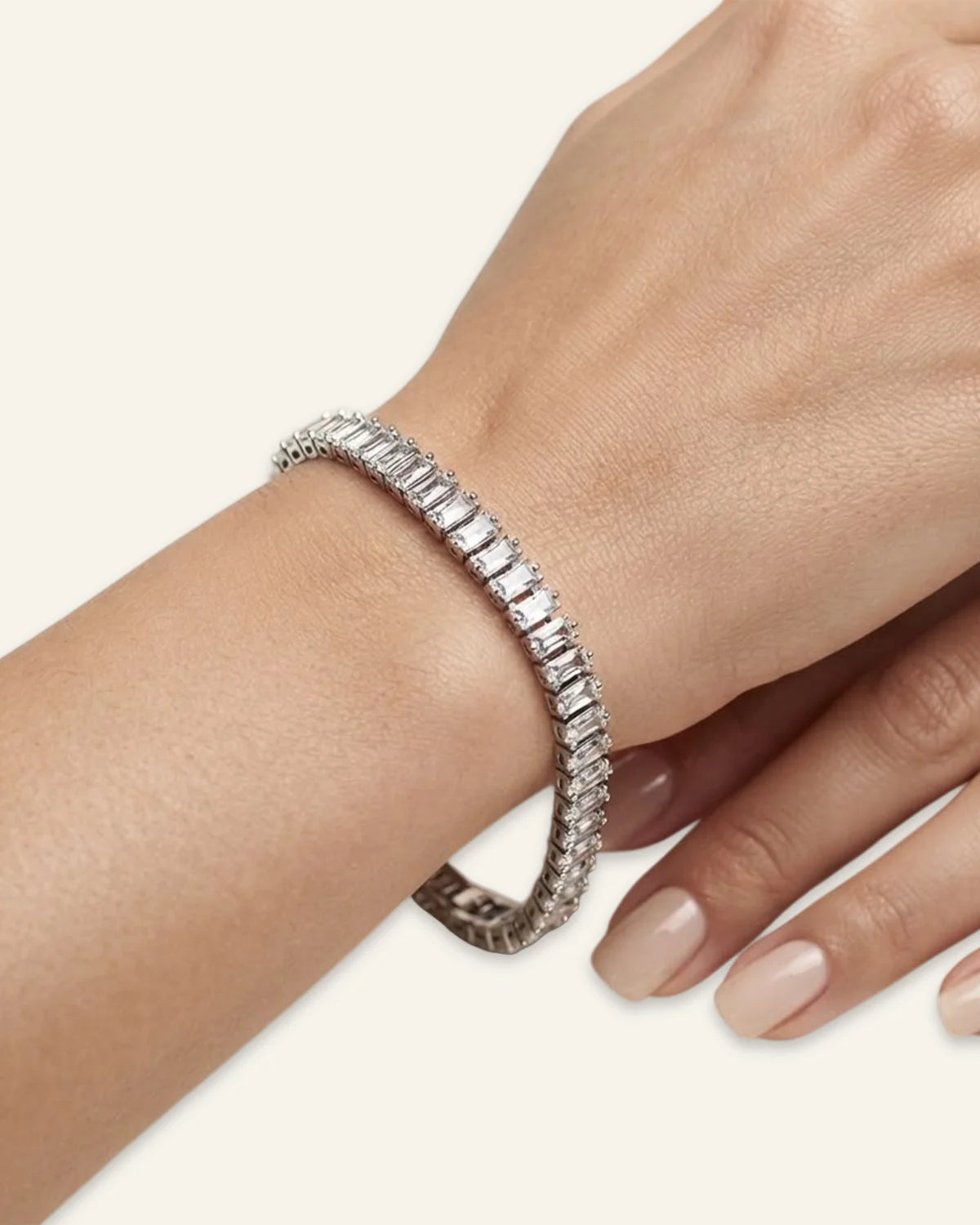 A close-up of a woman's wrist and hand wearing a silver dazzling baguette cut  tennis bracelet with rectangular clear stones, set against a solid light beige background.