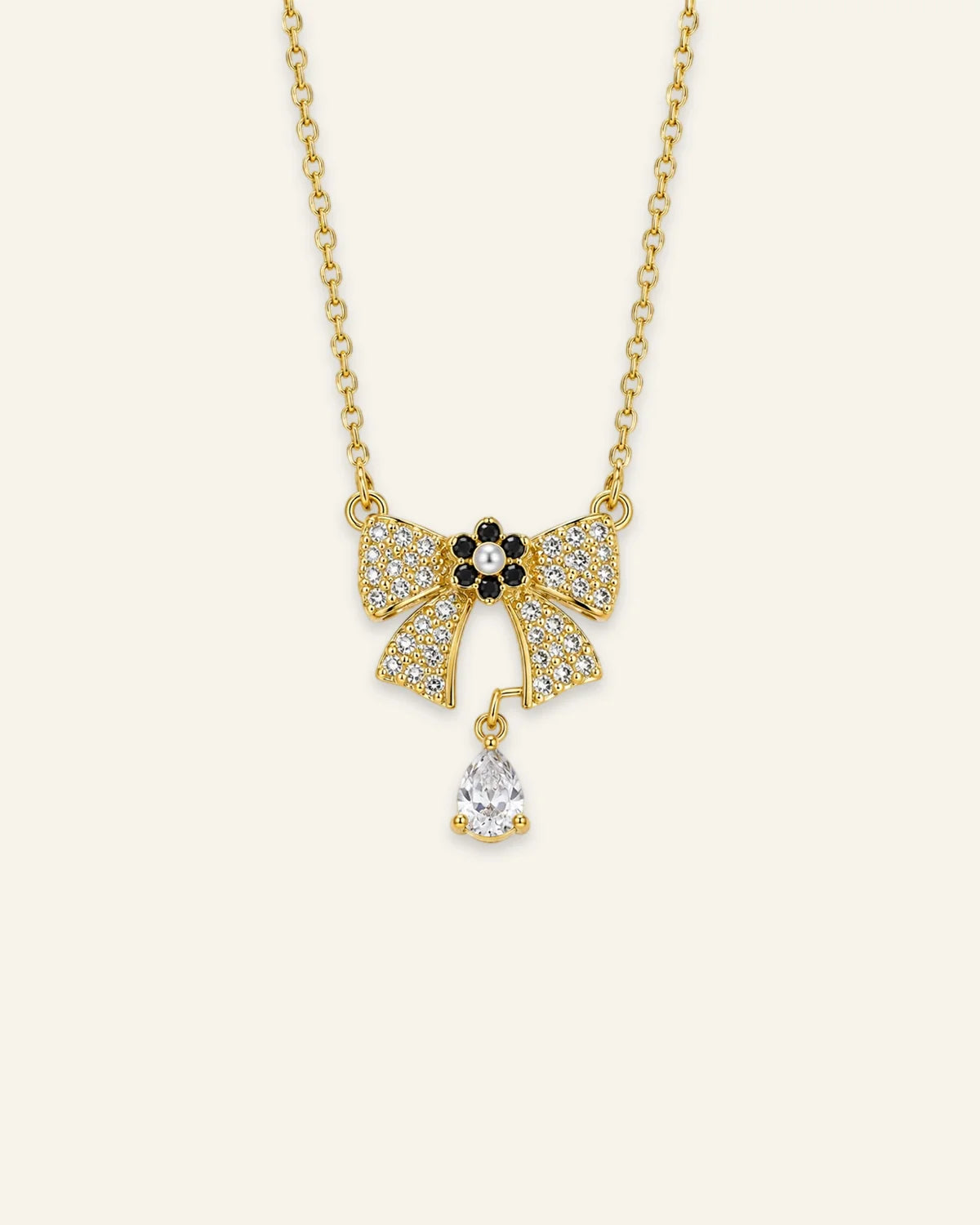 Gold chain necklace featuring a paved crystal ribbon bow pendant with a black-and-pearl flower centerpiece and a dangling teardrop-shaped gemstone.