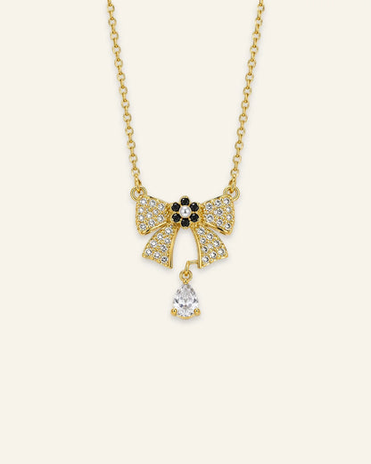 Gold chain necklace featuring a paved crystal ribbon bow pendant with a black-and-pearl flower centerpiece and a dangling teardrop-shaped gemstone.