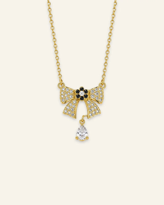 Gold chain necklace featuring a paved crystal ribbon bow pendant with a black-and-pearl flower centerpiece and a dangling teardrop-shaped gemstone.