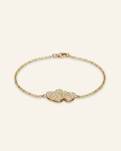 Heart Charm Bracelet, a delicate gold-tone chain bracelet featuring twin interlocking heart charms densely set with shining stones, displayed on a clean neutral background.