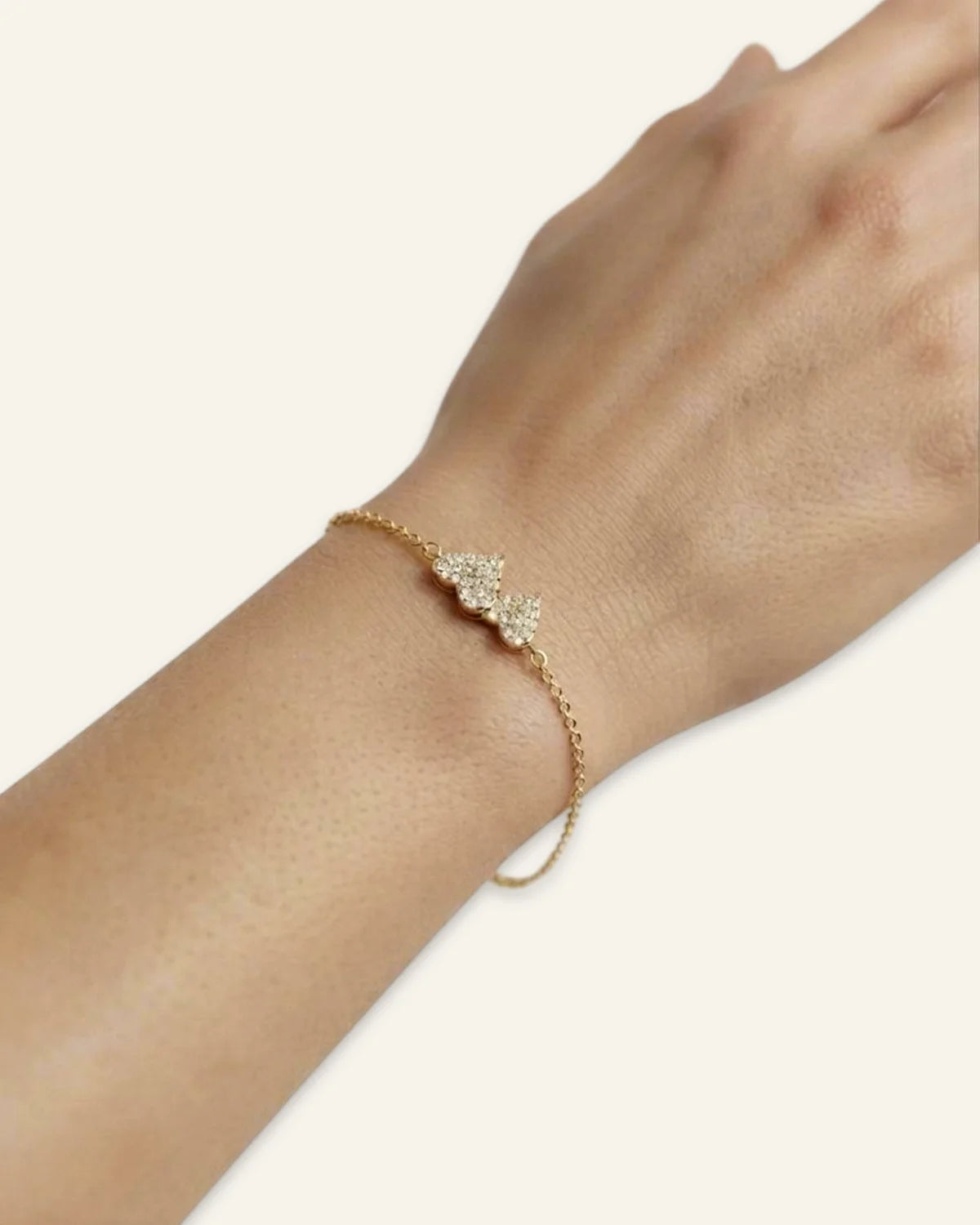Heart Charm Bracelet, a delicate gold-tone chain bracelet featuring twin interlocking heart charms densely set with shining stones, worn on a wrist  displayed on a clean neutral background.