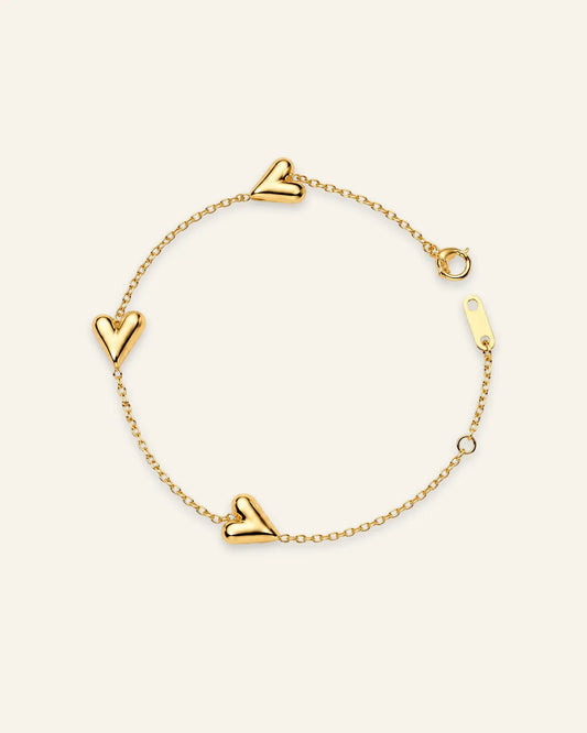 A delicate gold chain bracelet featuring three polished heart charms spaced along the chain, displayed on a clean, solid off-white background.
