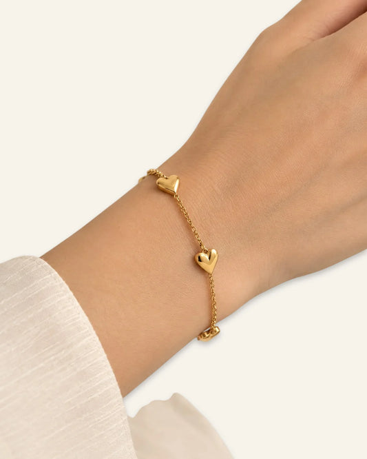 Heart Link Charm Bracelet, a delicate gold-tone chain bracelet worn on a wrist featuring  polished puffy heart charms along the band against a neutral background.