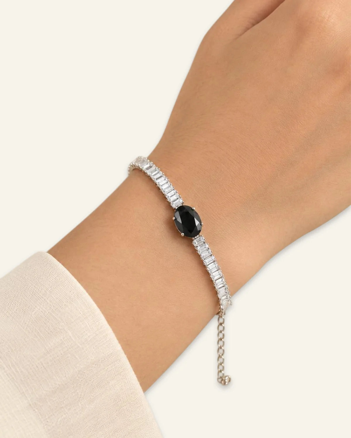 Midnight Luxe Bracelet, a silver-tone crystal line bracelet featuring a large central black oval gemstone flanked by rows of rectangular-cut clear stones  and an adjustable chain extension wore on a wrist of a hand on a neutral background.